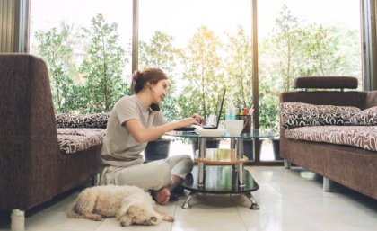 Image: Adobe. A woman sits on the floor in front of a low table using a phone and laptop computer, a dog is next to her.
